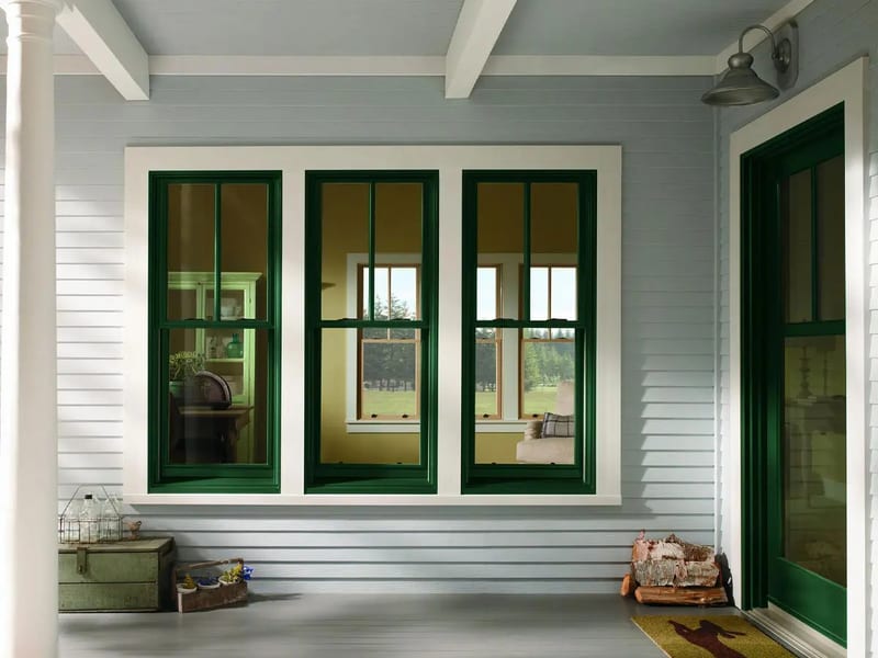 Three double-hung windows with green exteriors and colonial grilles in white lap siding