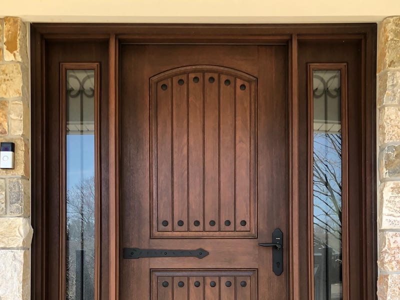 An arched timber-finish entry door with strap hinges and glazed sidelights in a stone surround
